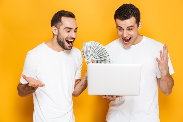 Deux amis joyeux et excités portant des t-shirts vierges isolés sur un mur jaune, utilisant un ordinateur portable, célébrant le succès, montrant des billets en argent