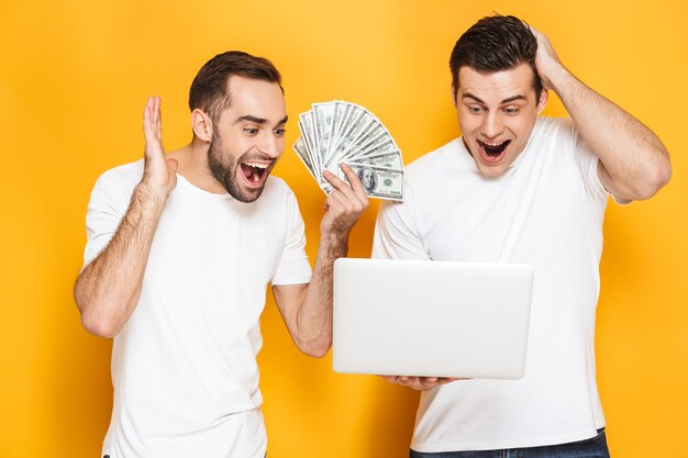 Deux amis joyeux et excités portant des t-shirts vierges isolés sur un mur jaune, utilisant un ordinateur portable, célébrant le succès, montrant des billets en argent