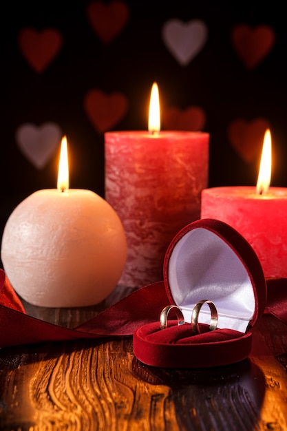 Photo deux anneaux de mariage dans une boîte cadeau rouge avec trois bougies flamme cire avec ruban dans une lumière romantique sombre sur les coeurs