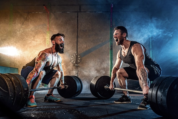 Deux Athlètes Tatoués Barbus Musclés S'entraînant, Soulevant Une Barre De Poids Lourde En Fumée Au Gymnase