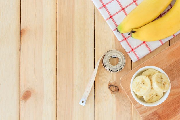 Deux bananes et tranches de banane dans un bol blanc avec un ruban à mesurer sur une table en bois