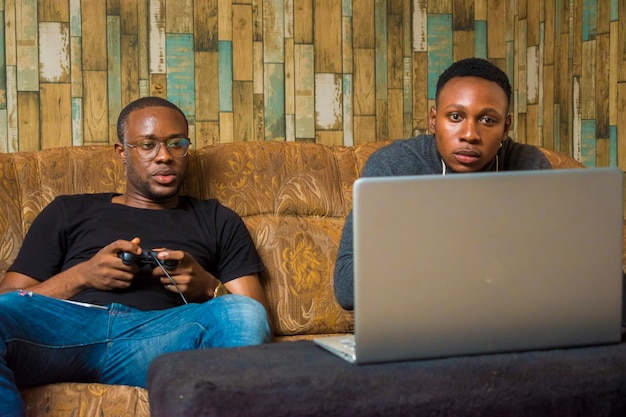 Deux beaux mecs africains assis sur le canapé jouant à des jeux vidéo avec manette de jeu, manette de jeu, pad