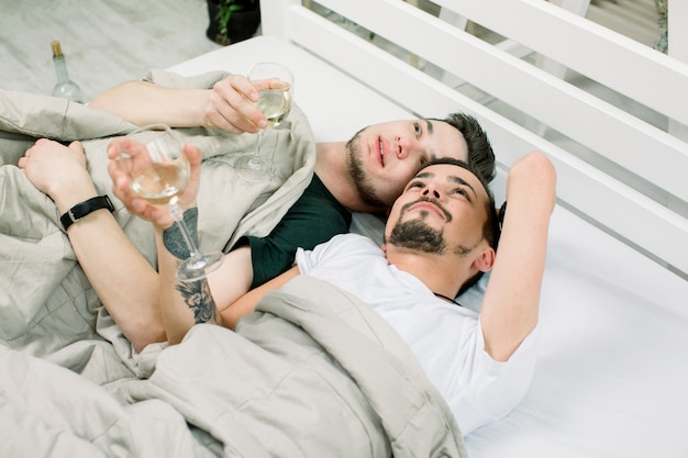 Deux beaux mecs boivent du vin blanc. allongé dans son lit à la maison. Jeune, gay, couple, mensonge, couverture, lit, lumière, salle