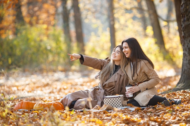 Deux belles amies passent du temps sur une couverture de pique-nique sur l'herbe. Deux jeunes soeurs souriantes faisant un pique-nique en mangeant un croissant dans le parc d'automne. Filles brunes et blondes portant des manteaux.