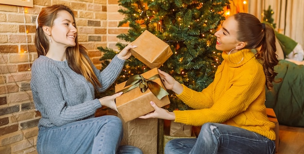 Deux belles copines joyeuses joyeuses jeunes filles donnent des cadeaux de Noël sur le fond d'un arbre du nouvel an à la maison