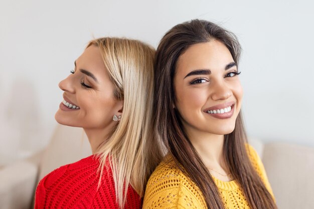 Deux belles femmes aux cheveux blonds et bruns portant des pulls tricotés assis sur le canapé dos à dos et souriant