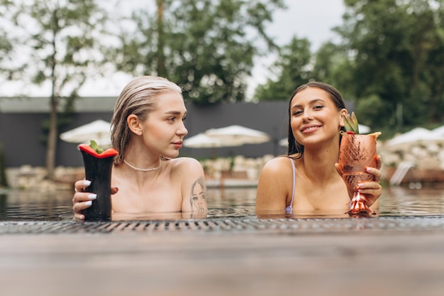 Deux belles femmes brune et blonde discutant en buvant des cocktails et se relaxant au bord de la piscine en été