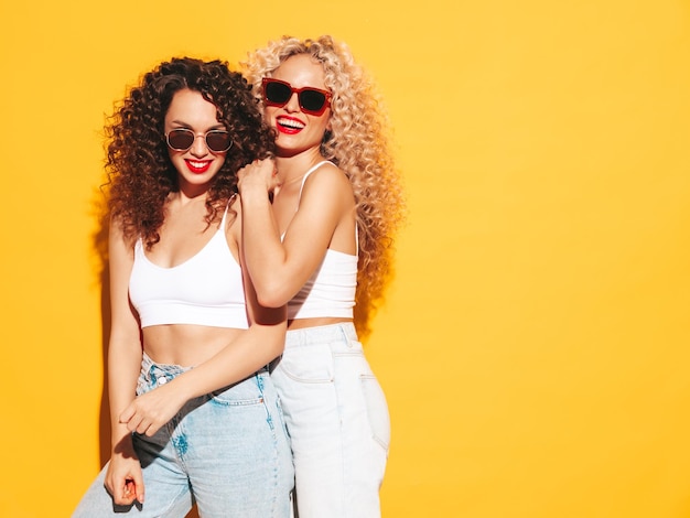 Deux belles femmes hipster souriantes sexy en maillot de bain d'été rouge maillots de bain Modèles à la mode avec une coiffure de boucles afro s'amusant en studio Femme chaude isolée sur jaune
