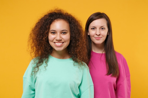 Deux belles femmes souriantes amies filles européennes et afro-américaines en vêtements verts roses posant isolées sur le portrait en studio de fond orange jaune. Concept de style de vie des gens. Maquette de l'espace de copie.