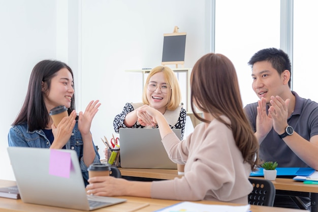 Deux belles filles asiatiques se serrant la main et se regardant avec le sourire pendant que leurs collègues applaudissent à la table du bureau