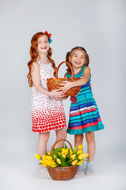 Deux belles filles se tiennent près du panier avec des tulipes, tenant un panier avec des oeufs de Pâques dans leurs mains et riant drôlement. Fond blanc. Photo drôle.