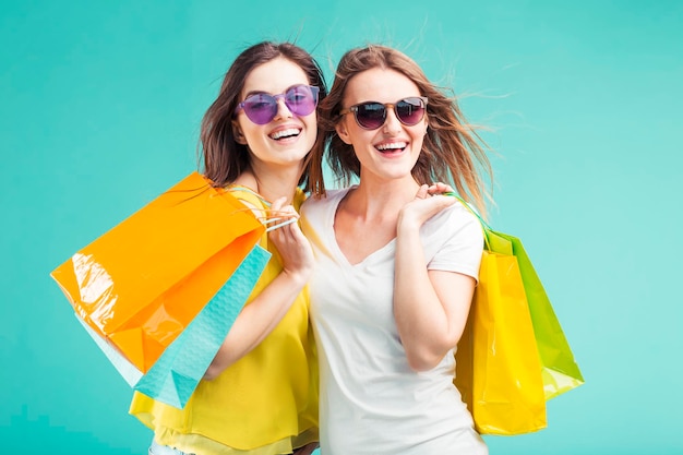 Deux belles filles souriantes accros du shopping portent des lunettes de soleil et contiennent beaucoup de sacs de shopping colorés sur fond bleu