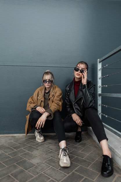 Deux belles jeunes adolescentes vêtues de vêtements urbains à la mode avec des jeans et des baskets sont assises près d'un mur métallique