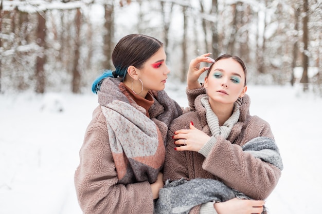 Deux belles jeunes copines à la mode à la mode avec un maquillage coloré créatif dans des vêtements élégants d'hiver pose dans un parc d'hiver avec de la neige