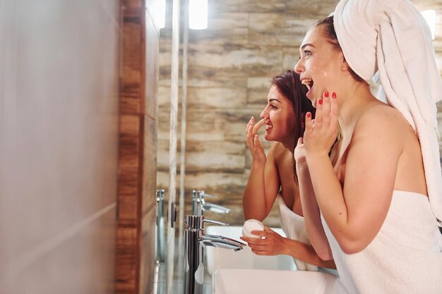 Deux belles jeunes femmes debout dans la salle de bain près du miroir ensemble et se nettoyant le visage.