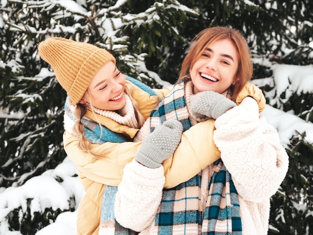 Deux belles jeunes femmes hipster souriantes dans des vêtements chauds à la mode et des écharpes. Femmes insouciantes posant dans la rue dans le parc. Des modèles purs positifs s'amusant dans la neige. Profiter des moments d'hiver. Notion de Noël