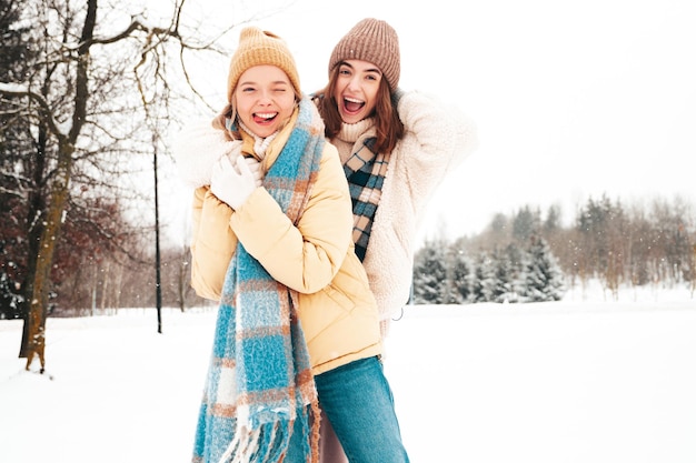 Deux belles jeunes femmes hipster souriantes dans des vêtements chauds à la mode et des écharpes. Femmes insouciantes posant dans la rue dans le parc. Des modèles purs positifs s'amusant dans la neige. Profiter des moments d'hiver. Notion de Noël