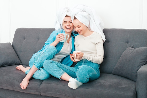 Deux belles jeunes femmes souriantes assises sur le canapé. modèles insouciants posant à l'intérieur dans un appartement chic ou une chambre d'hôtel. Ils font des soins de beauté à la maison