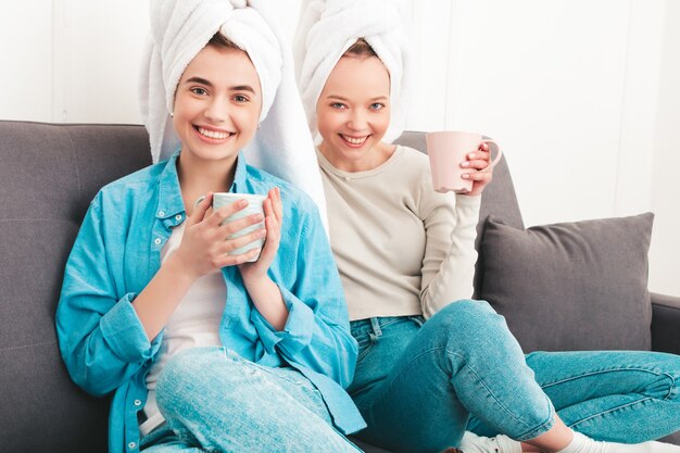 Deux belles jeunes femmes souriantes assises sur le canapé. modèles insouciants posant à l'intérieur dans un appartement chic ou une chambre d'hôtel. Ils font des soins de beauté à la maison