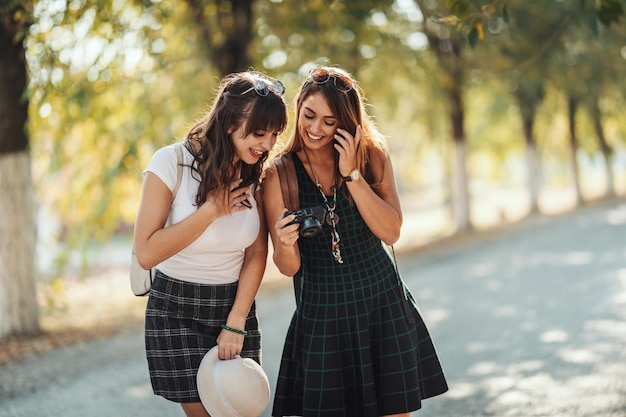 Deux belles jeunes femmes souriantes avec des sacs à dos sur le dos regardent les photos sur l'appareil photo pendant qu'elles marchent le long de l'avenue ensoleillée d'automne.