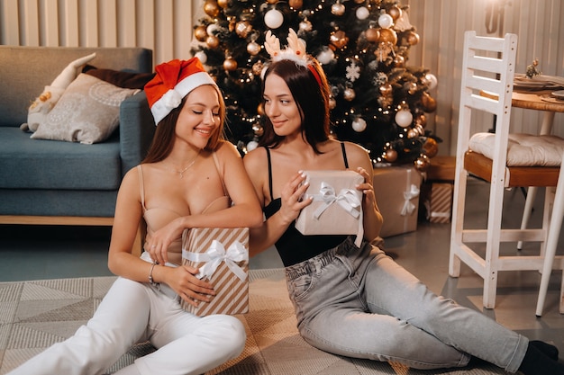 Deux belles jeunes filles avec des cadeaux de Noël dans leurs mains sont assises près de l'arbre de Noël dans la maison.amis avec des robes de Noël et des cadeaux emballés