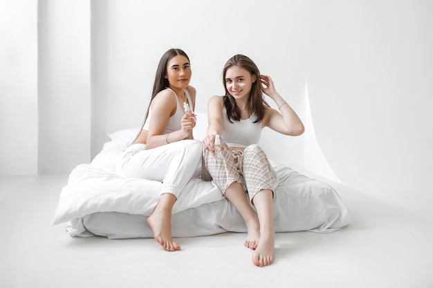 Deux belles jeunes filles avec des cosmétiques sont assises sur un lit blanc Les femmes utilisent de la crème sérique et de la lotion pour le visage pour prendre soin de leur peau Les copines du spa à domicile se reposent et se détendent