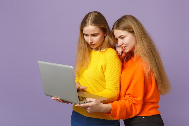 Deux belles jeunes filles jumelles blondes dans des vêtements colorés vifs tiennent, à l'aide d'un ordinateur portable isolé sur un mur bleu violet pastel. Concept de mode de vie familial de personnes. .