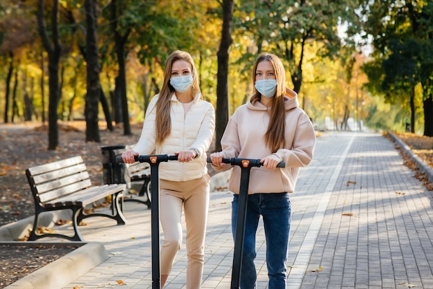 Deux belles jeunes filles masquées montent des scooters électriques dans le parc par une chaude journée d'automne.