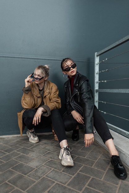 Deux belles jeunes filles sœurs en streetwear à la mode avec veste, lunettes, jisami et baskets sont assises près d'un mur sombre en métal