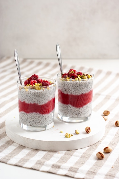 Deux bocal en verre de pudding de chia avec framboise, pistache et confiture rouge sur une table blanche avec nappe à rayures.