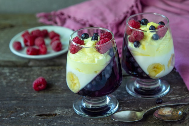 Deux bocaux en verre avec dessert froid (panna cotta, gelée, mousse, pudding) avec des couches de myrtille violette et de banane blanche sur table rustique en bois