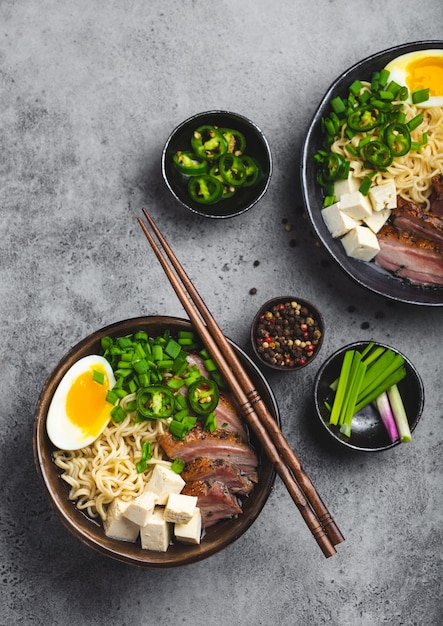 Deux bols de savoureuse soupe de nouilles asiatiques ramen avec bouillon de viande, tofu, porc, œuf avec jaune sur fond de béton rustique gris, gros plan, vue de dessus. Soupe de ramen japonaise savoureuse et chaude pour le dîner à l'asiatique