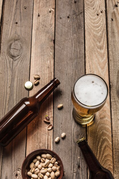 Deux bouteilles de bière brune, un verre de bière et de mousse et une assiette de pistaches sur un fond en bois.