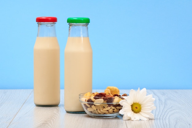 Deux bouteilles de lait frais avec bouchon rouge et vert, bol en verre avec muesli et fleur de camomille sur fond bleu