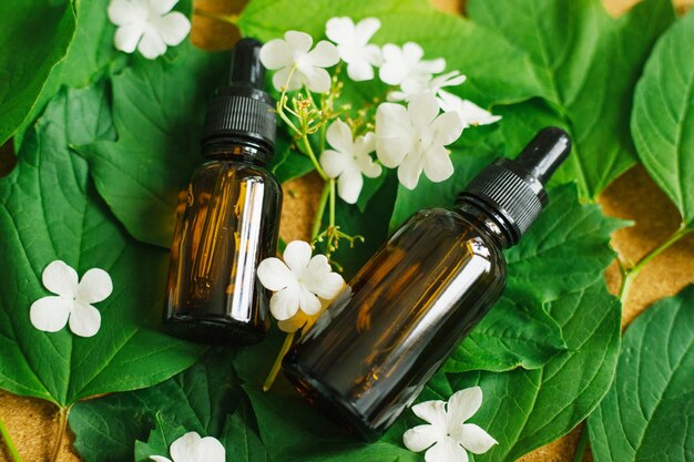 Deux Bouteilles En Verre D'huile Cosmétique Se Trouvent Sur Un Fond De Feuilles Vertes Et De Fleurs Blanches.