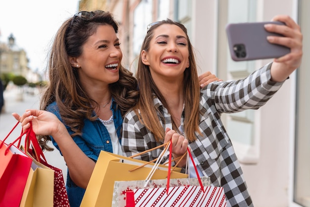 Deux charmantes femmes souriantes prenant un selfie sur un smartphone tenant des sacs à provisions