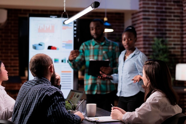 Deux collègues discutant d'idées commerciales avec des membres du conseil d'administration dans un bureau de démarrage travaillant des heures supplémentaires lors d'une réunion de fin de soirée. Des collègues de travail afro-américains réfléchissent à une stratégie marketing avec l'équipe de vente.