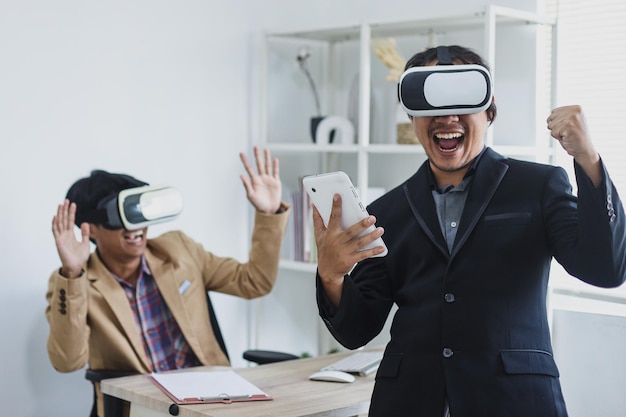 Deux collègues masculins asiatiques excités en costume utilisant des lunettes VR jouant à des jeux pendant les pauses au bureau