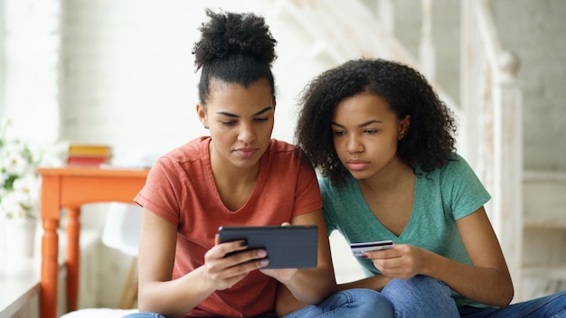 Deux copines bouclées de race mixte joyeuses faisant du shopping en ligne avec une tablette