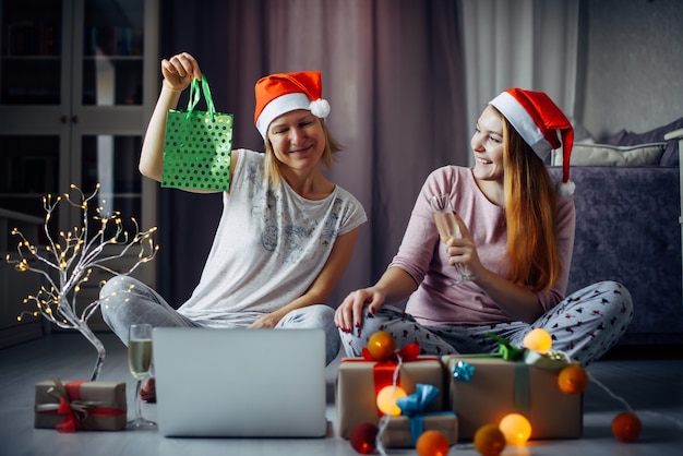 Deux copines gaies ouvrent des coffrets cadeaux de Noël lors d'une vidéoconférence en ligne avec la famille sur un ordinateur portable. Les sœurs échangent des cadeaux à l'intérieur de la maison avec des décorations festives.