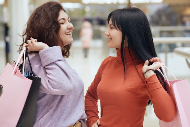 Deux copines heureuses qui se sourient en se tenant debout avec des sacs à provisions dans le centre commercial, des dames portant des vêtements décontractés, de bonne humeur, appréciant d'acheter de nouveaux vêtements.