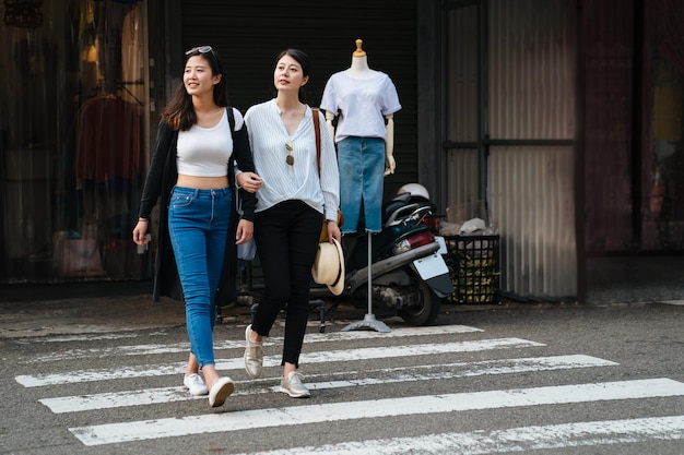 deux copines millénaires se tenant la main et riant à travers le passage pour piétons. pleine longueur de charmantes sœurs asiatiques regardant du même côté vérifiant avec les voitures et la circulation marchant sur la route zébrée à l'extérieur.