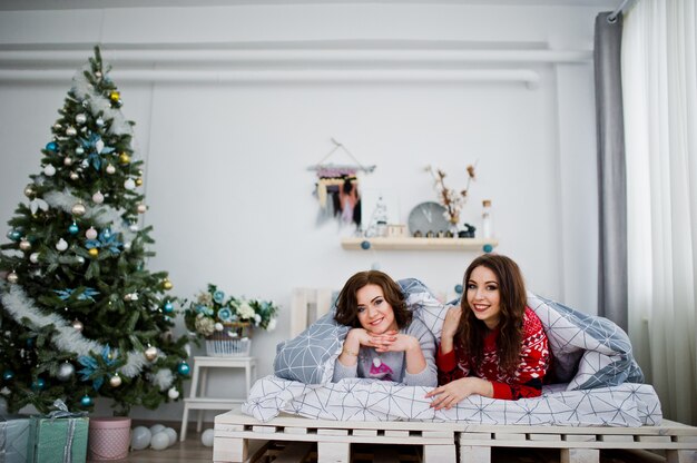 Deux copines portent des chandails d’hiver s’amusant sur un lit dans une chambre avec des décorations de Noël.