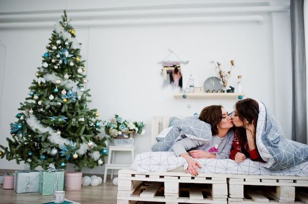 Deux copines portent des chandails d’hiver s’amusant sur un lit dans une chambre avec des décorations de Noël.