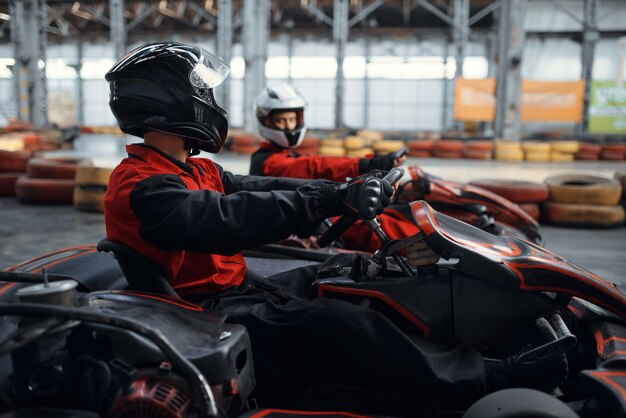 Deux coureurs de kart se battent pour la victoire sur les genoux, vue de côté, karting auto sport indoor. Course de vitesse sur piste de karting fermée avec barrière de pneu. Compétition de véhicules rapide, loisirs à forte adrénaline