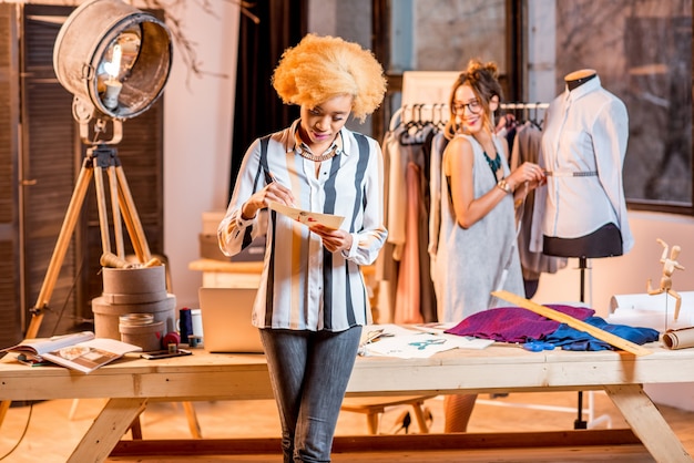 Photo deux créatrices de mode multiethniques travaillant avec des dessins de vêtements debout au bureau avec différents outils de couture et mannequin