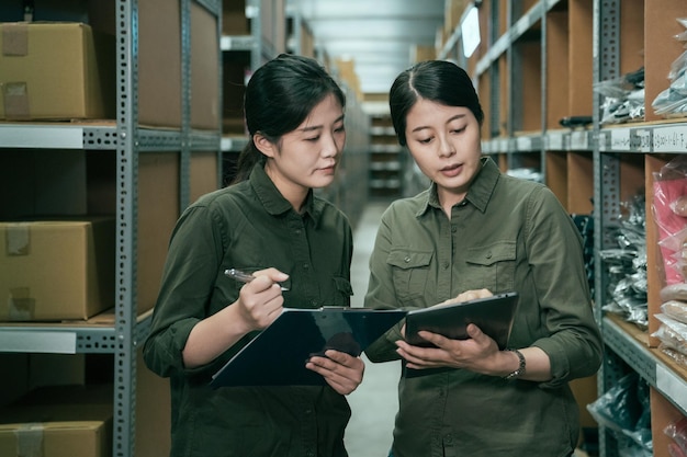 deux employées d'entrepôt de femme asiatique avec tablette et presse-papiers à l'intérieur. jeunes filles employées en uniforme discutant de la quantité de marchandises. personnel féminin debout entre une boîte en carton et des produits sur une étagère