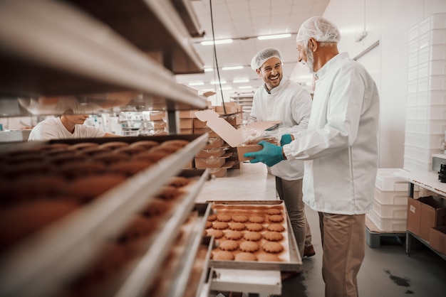 Deux employés caucasiens dévoués et assidus vêtus d'uniformes blancs stériles collectent et emballent des biscuits dans des boîtes. Intérieur de l'usine alimentaire.