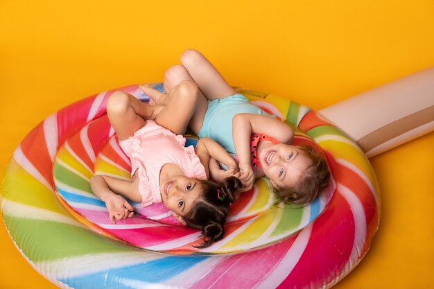 Deux enfant fille en maillot de bain allongé s'amusant sur une sucette de matelas gonflable coloré sur fond jaune.