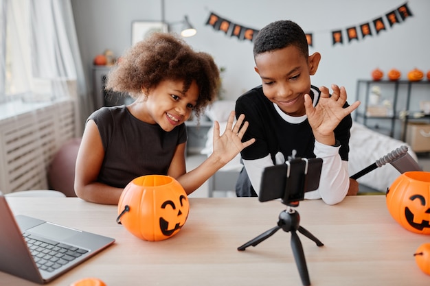 Deux enfants afro-américains souriants saluant un smartphone tout en discutant en vidéo en direct dans le hall...